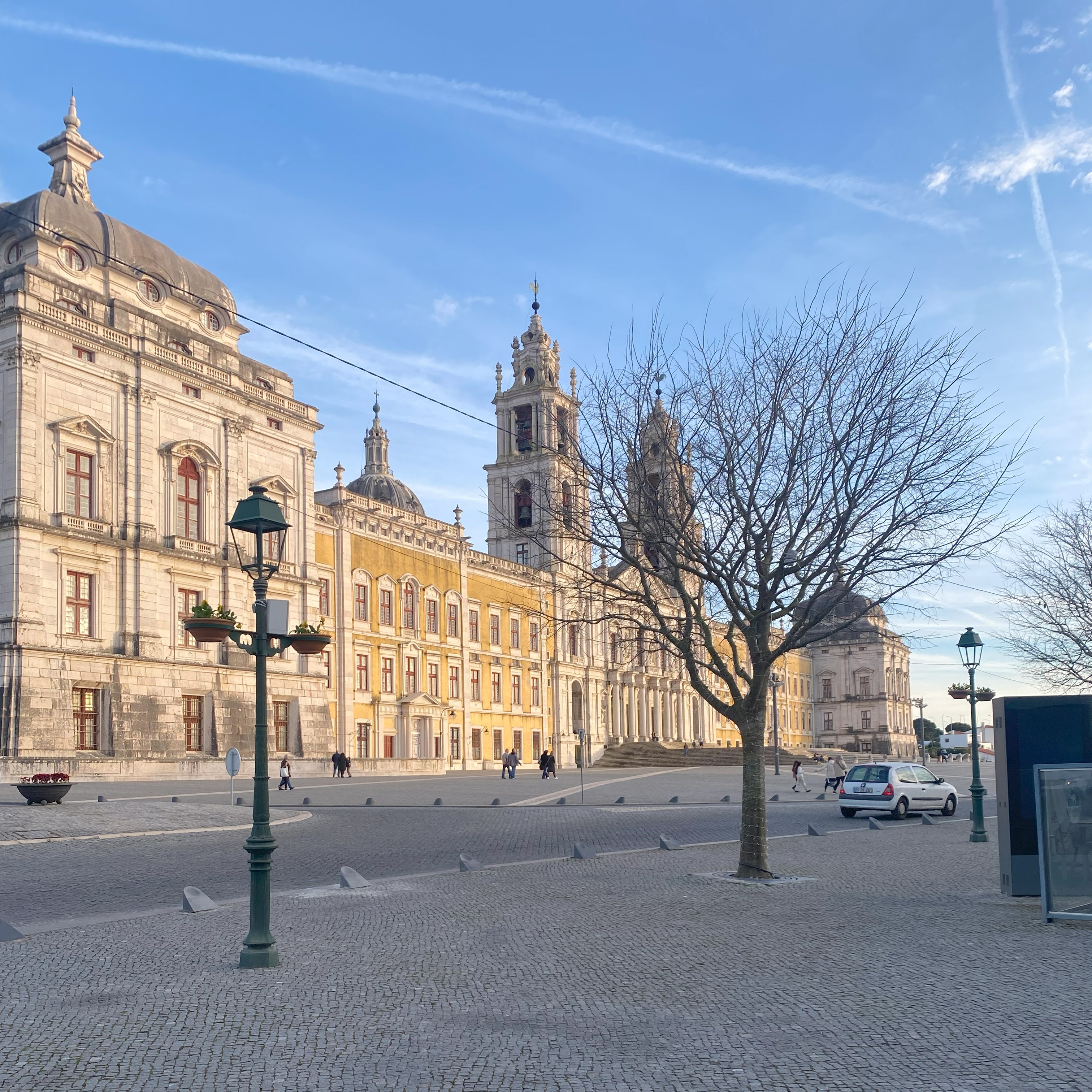 Praça de paralelepípedos em Mafra, Portugal, com o majestoso Palácio Nacional de Mafra ao fundo, sob um céu azul. Uma árvore seca e um poste de luz decorado se destacam em primeiro plano.